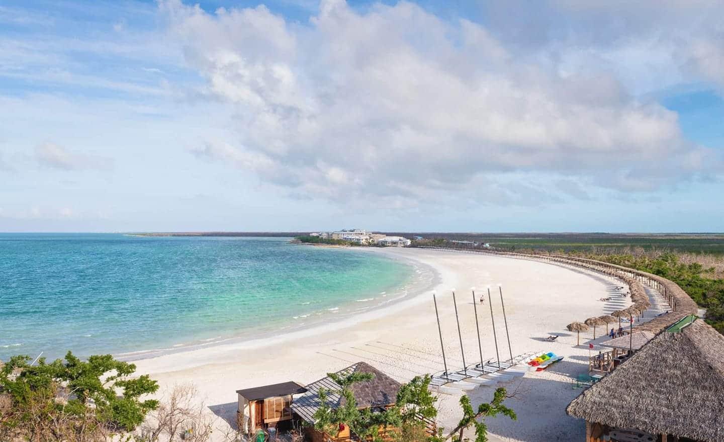 Aerial view of a curved white sand beach with turquoise water, surrounded by greenery and thatched-roof structures under a partly cloudy sky.