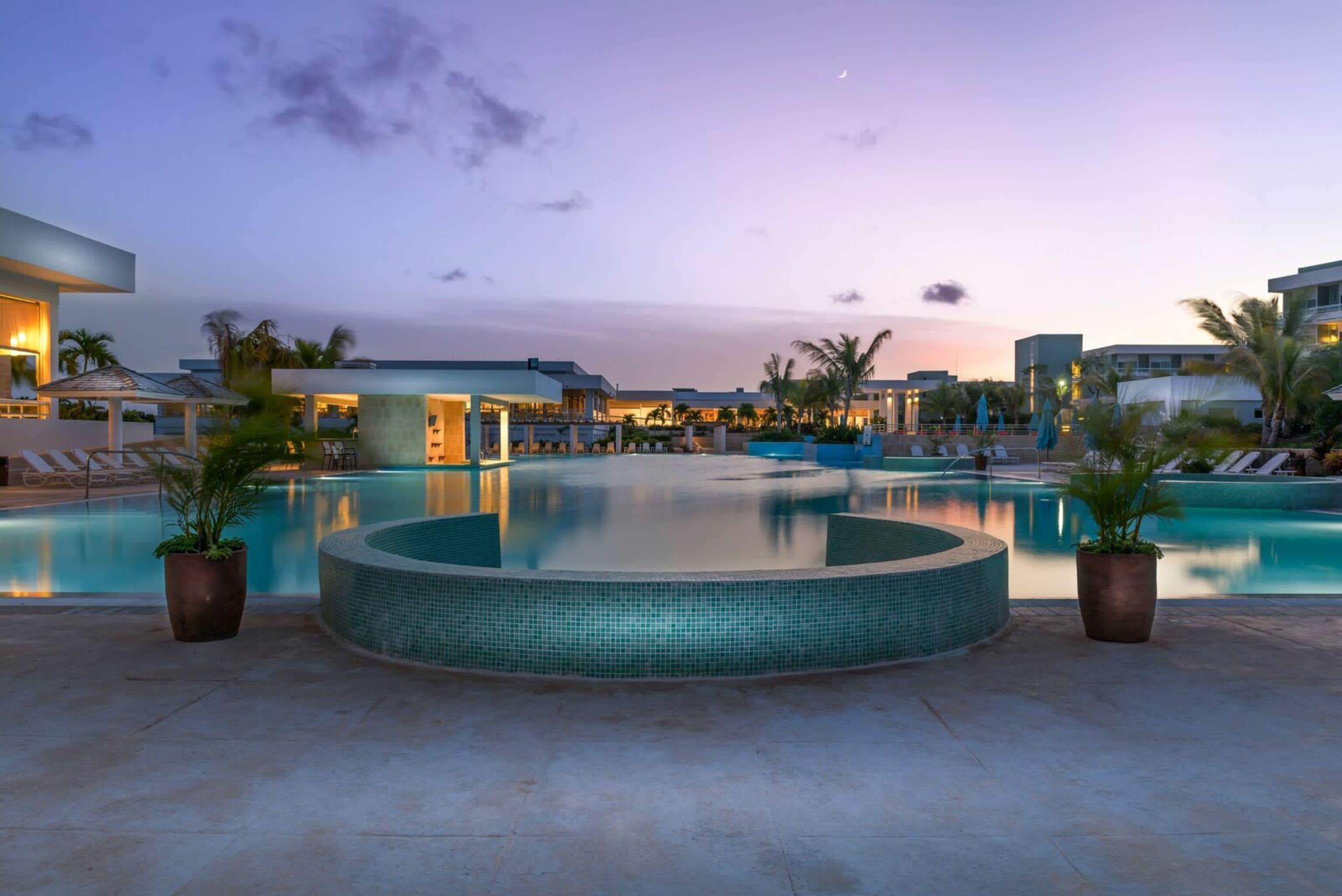 Twilight view of a serene resort pool with modern architecture, palm trees, and a crescent moon.