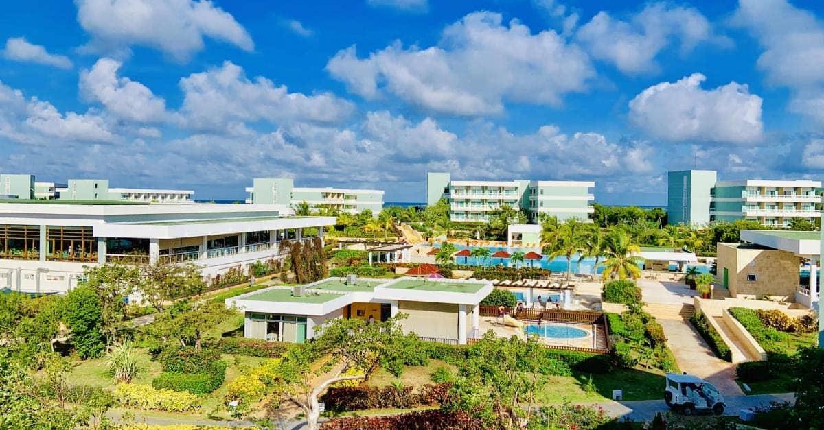 Aerial view of a resort with multiple buildings, a pool area, and lush greenery under a blue sky with clouds.